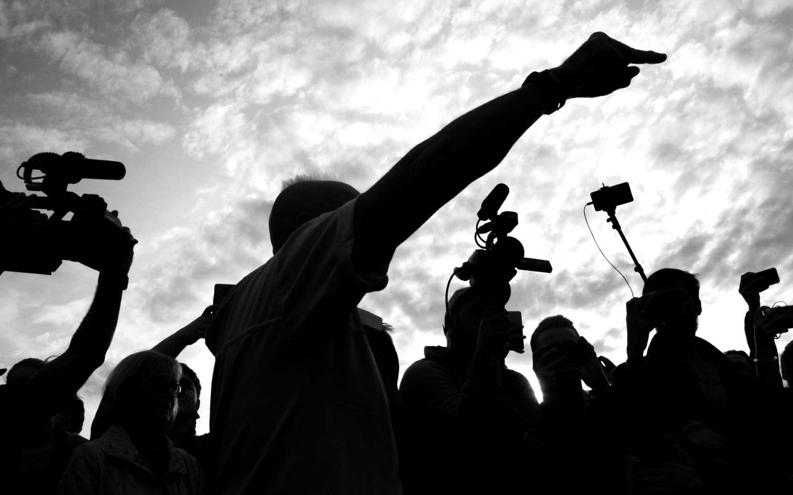 Silhouetted man points skyward while surrounded by cameras and microphones from the press