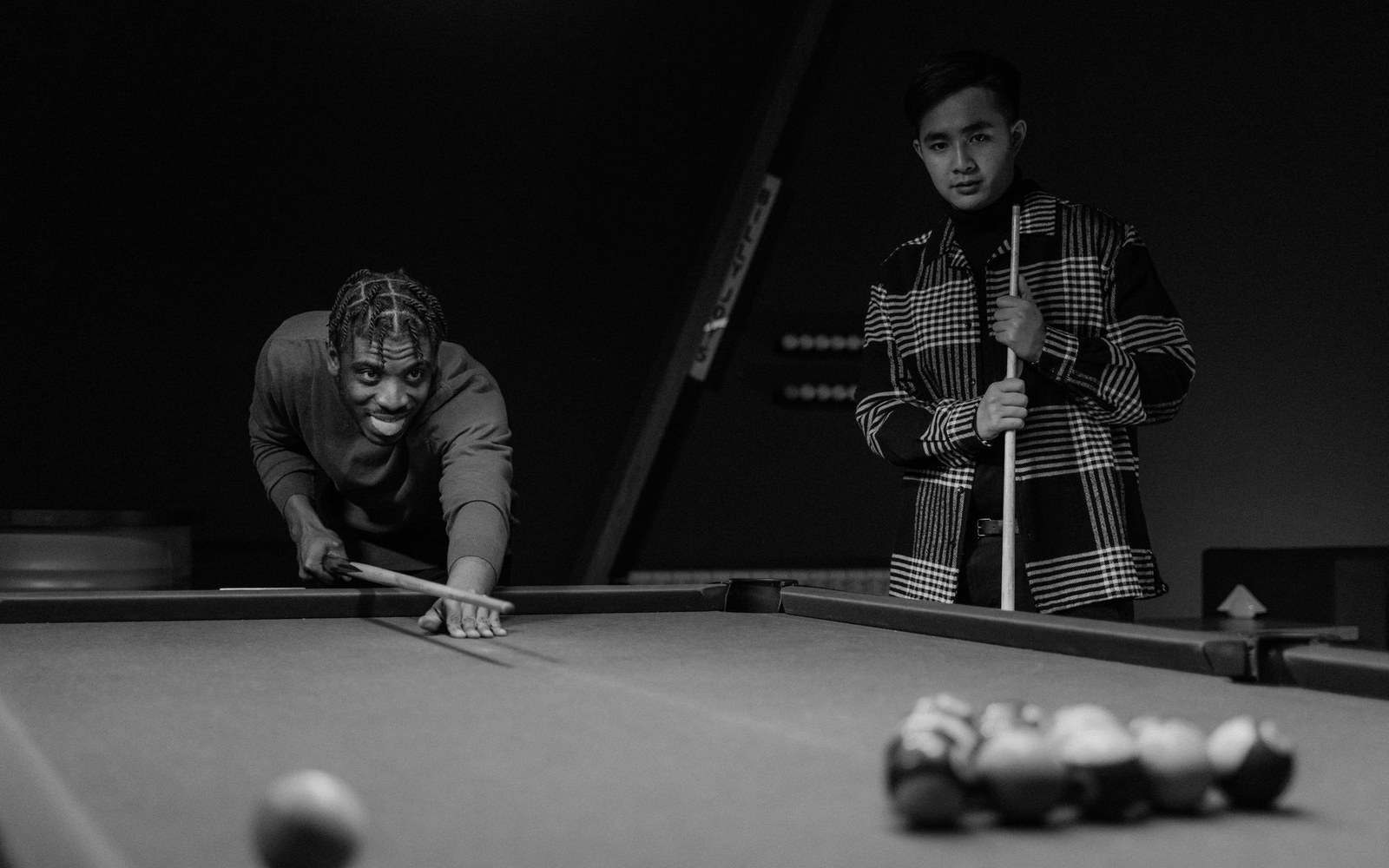 Two men play pool, one leaning to take a shot while the other watches with cue in hand