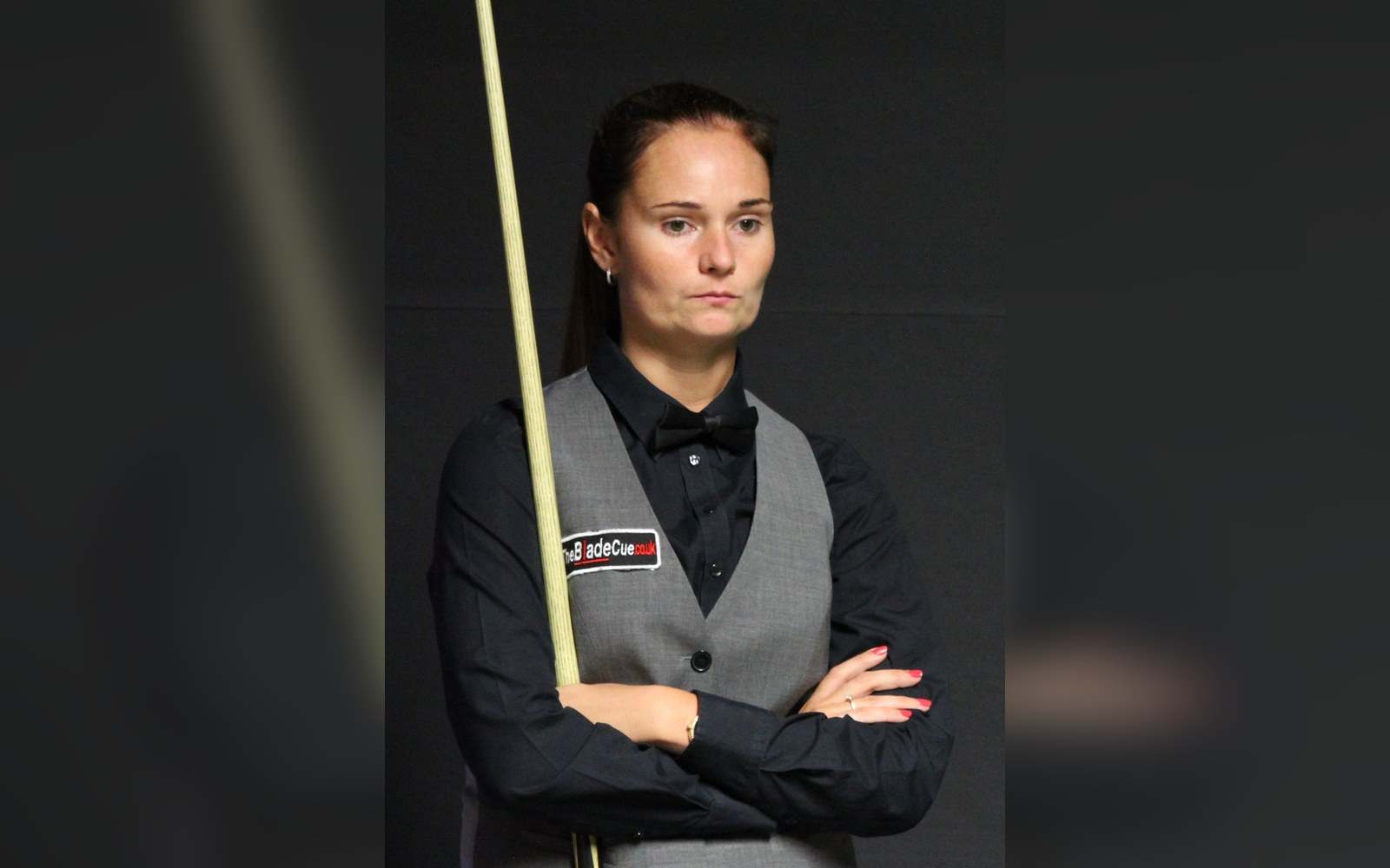 Snooker player in waistcoat and bow tie stands with arms crossed, holding a cue