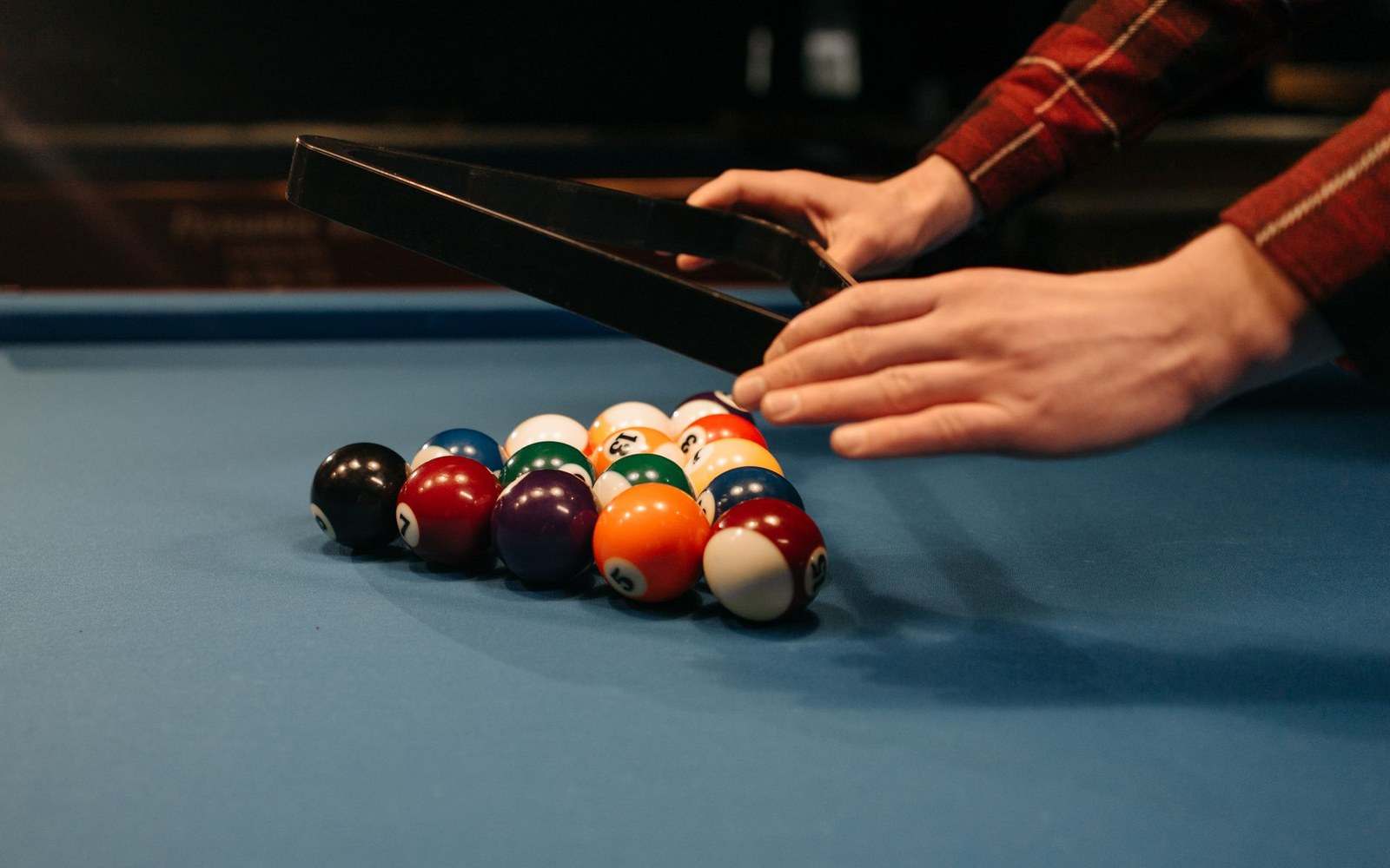 Hands lifting a triangle rack from freshly arranged billiard balls on a blue table